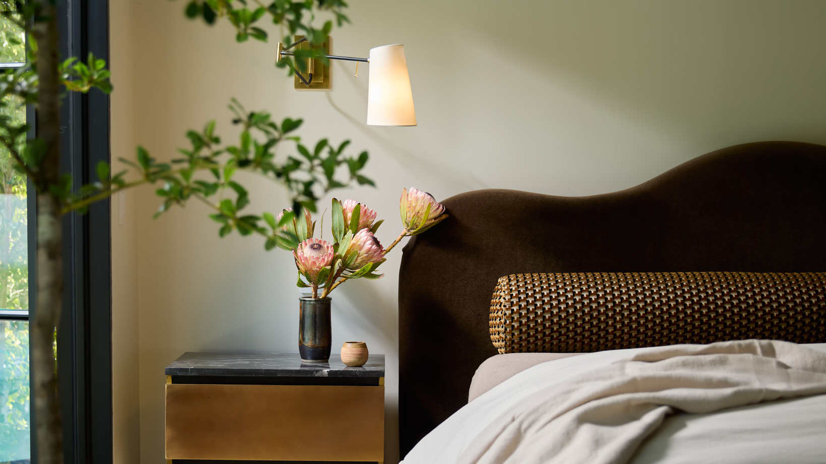 wavy brown headboard of a bed against a pale off-white wall with flowers on the nightstand next to it