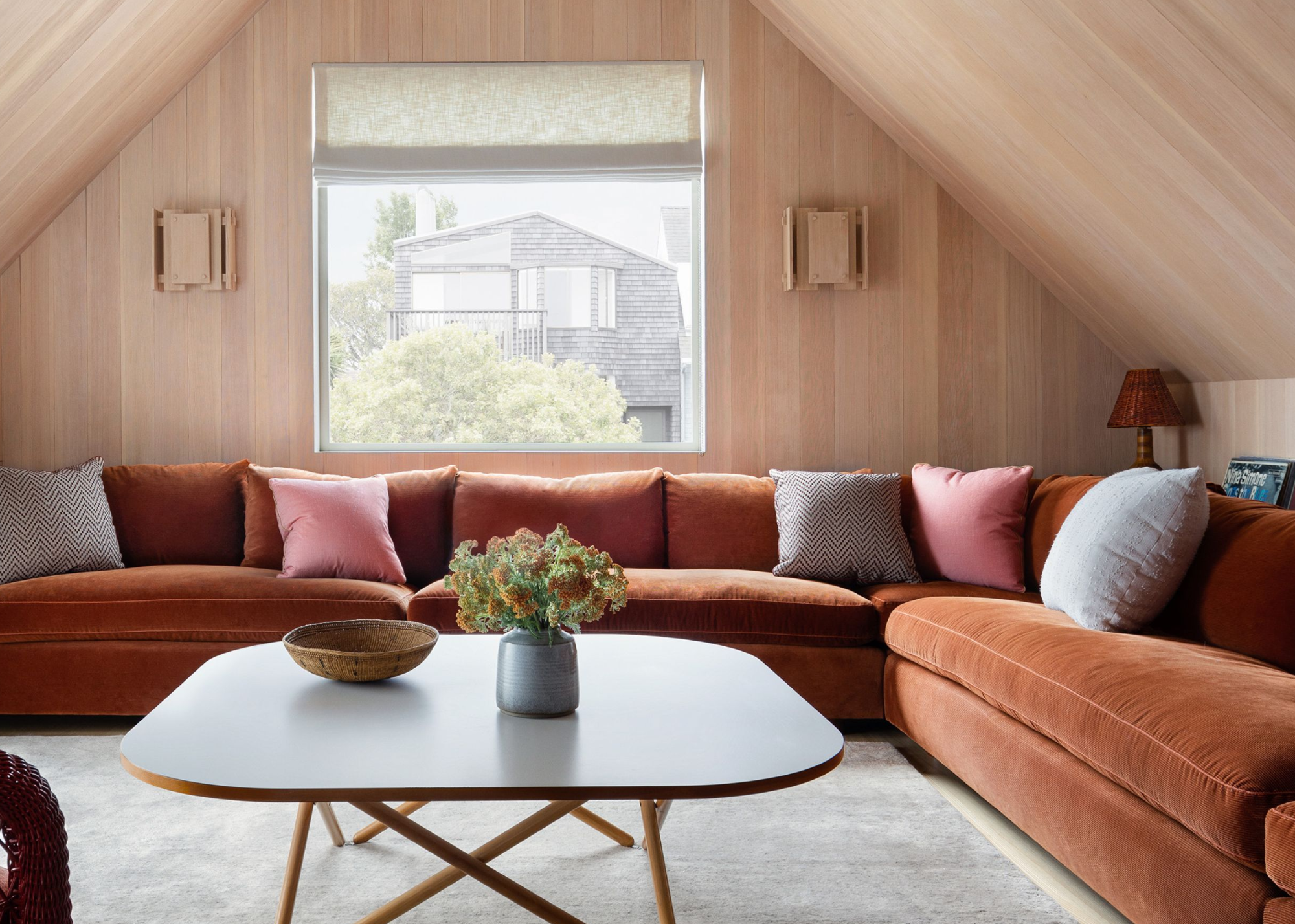 conversation room in attic with timber panelled walls and ceiling, rust-colored corner sofa with cushions, square coffee table with timber legs, beige rug, and wicker armchair