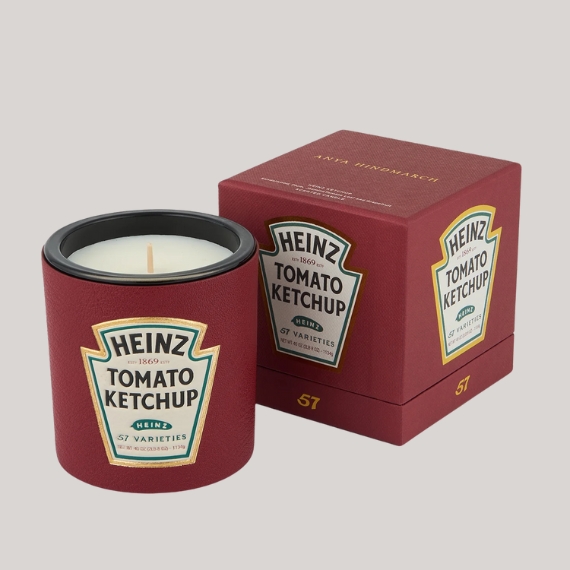 A ketchup tub-looking candle with a label "HEINZ TOMATO KETCHUP" sits next to its box in a similar style against a pale faint grey.
