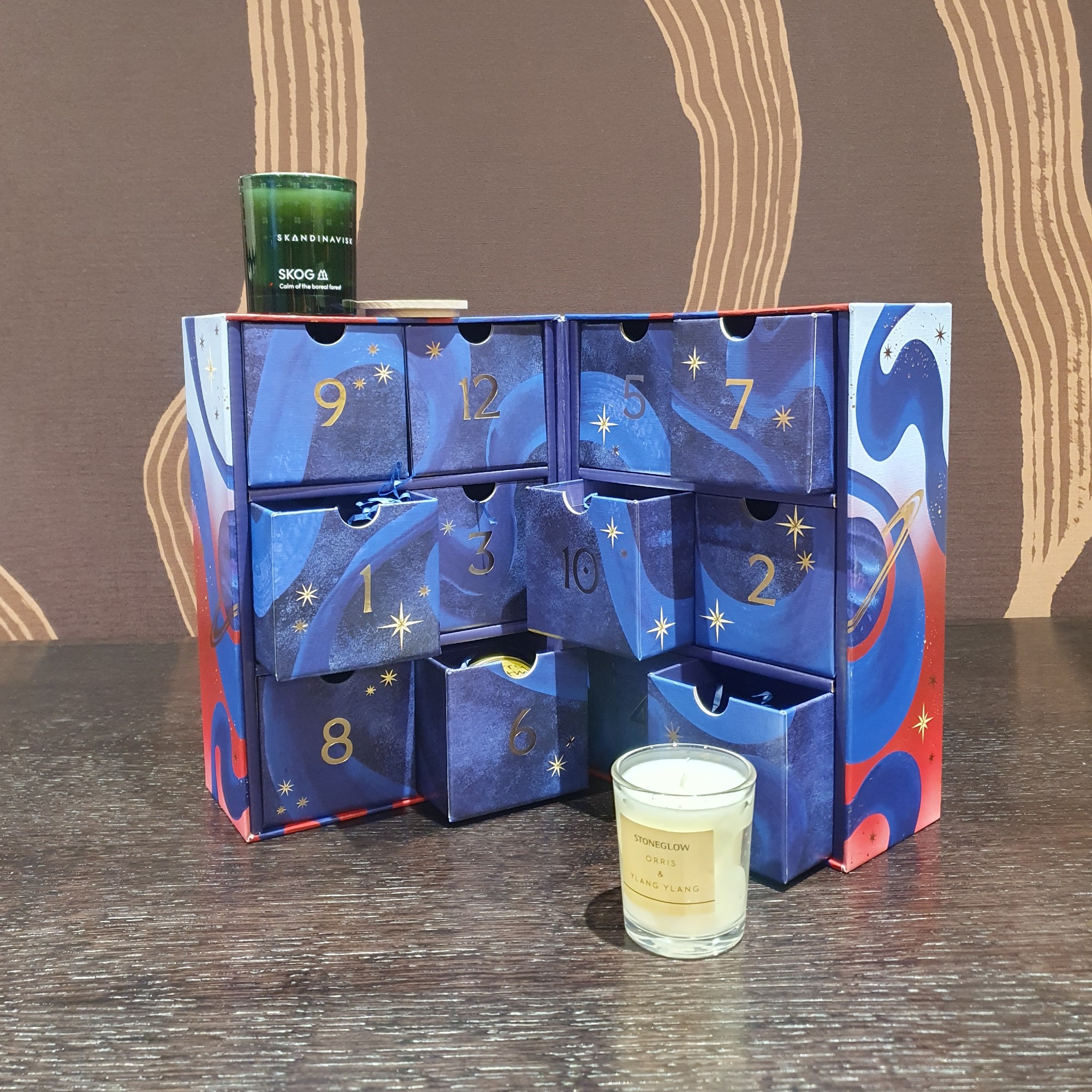 Open John Lewis scented candle advent calendar on a dark wood table with a brown wallpaper behind with two of the scented candles taken out 