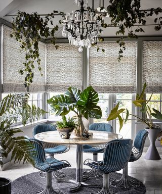 Biophilic dining scheme with chrome contrast dining table and chairs, palm plants, ivy, and ferns, and patterned window blinds.