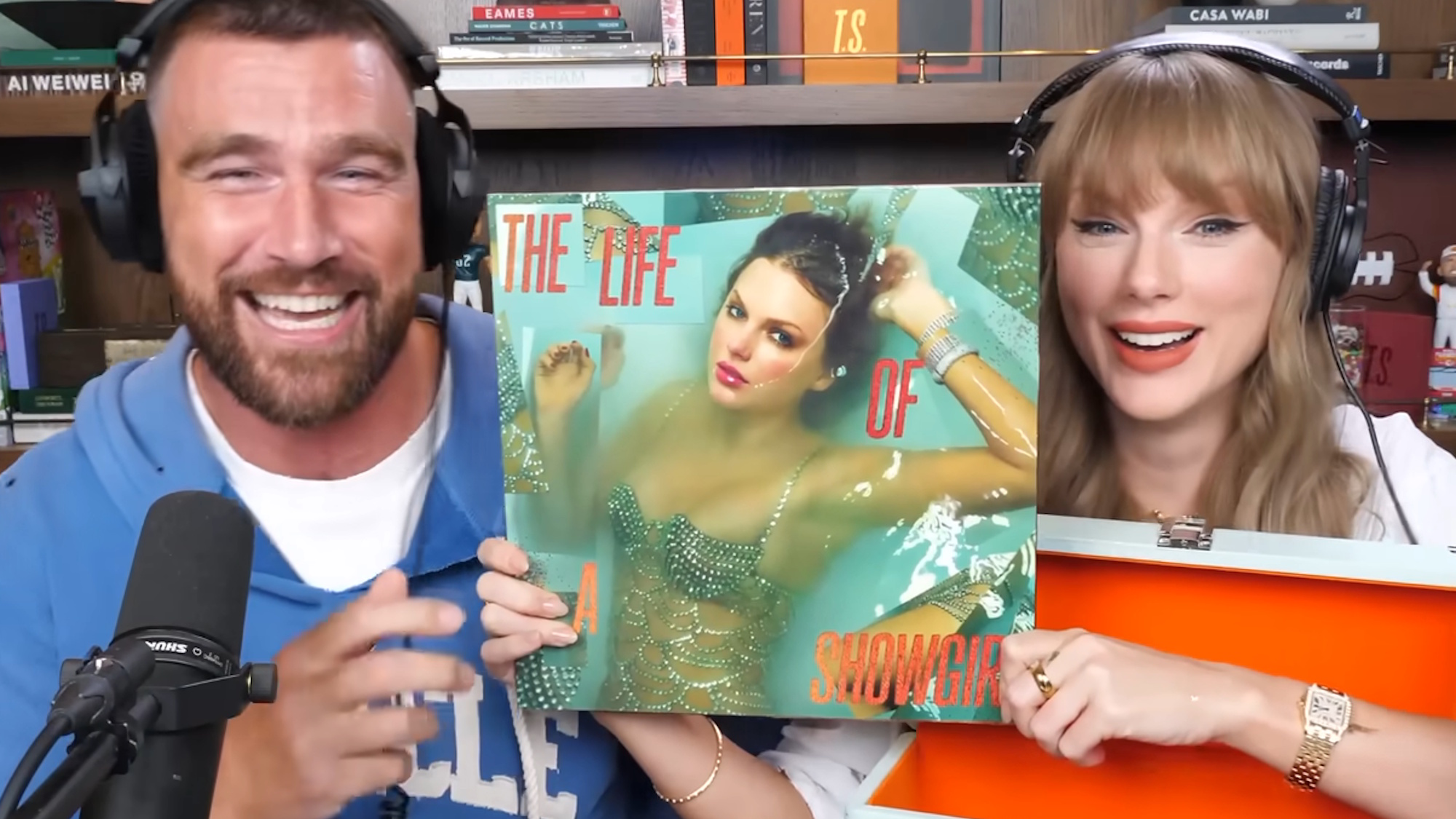 Travis Kelce and Taylor Swift smile as they show The Life of a Showgirl album cover art on the New Heights podcast.