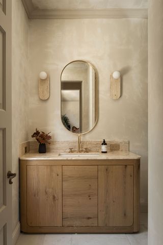 A beige bathroom with a wooden vanity unit and limewash walls with wall sconces either side of the oval mirror