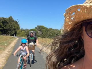 Family cycling holiday on Île de Ré shows the side of a female, with small child and man behind riding a paved cycle path with blue sky in the background