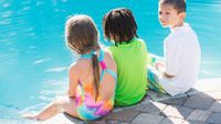 Picture of three children (a girl on the left-hand side and two boys on her right) sitting by the edge of a swimming pool. The boy who is furthest on the right-hand side has his head turned back and is looking into the distance.