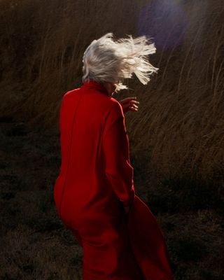 An elderly woman facing away from the camera wearing a red coat standing in front of some plants.