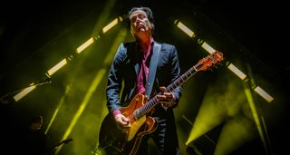 Troy Van Leeuwen of Queens of the Stone Age plays a red/orange Gretsch onstage, and is framed by a triangle of yellow-green stagelights.