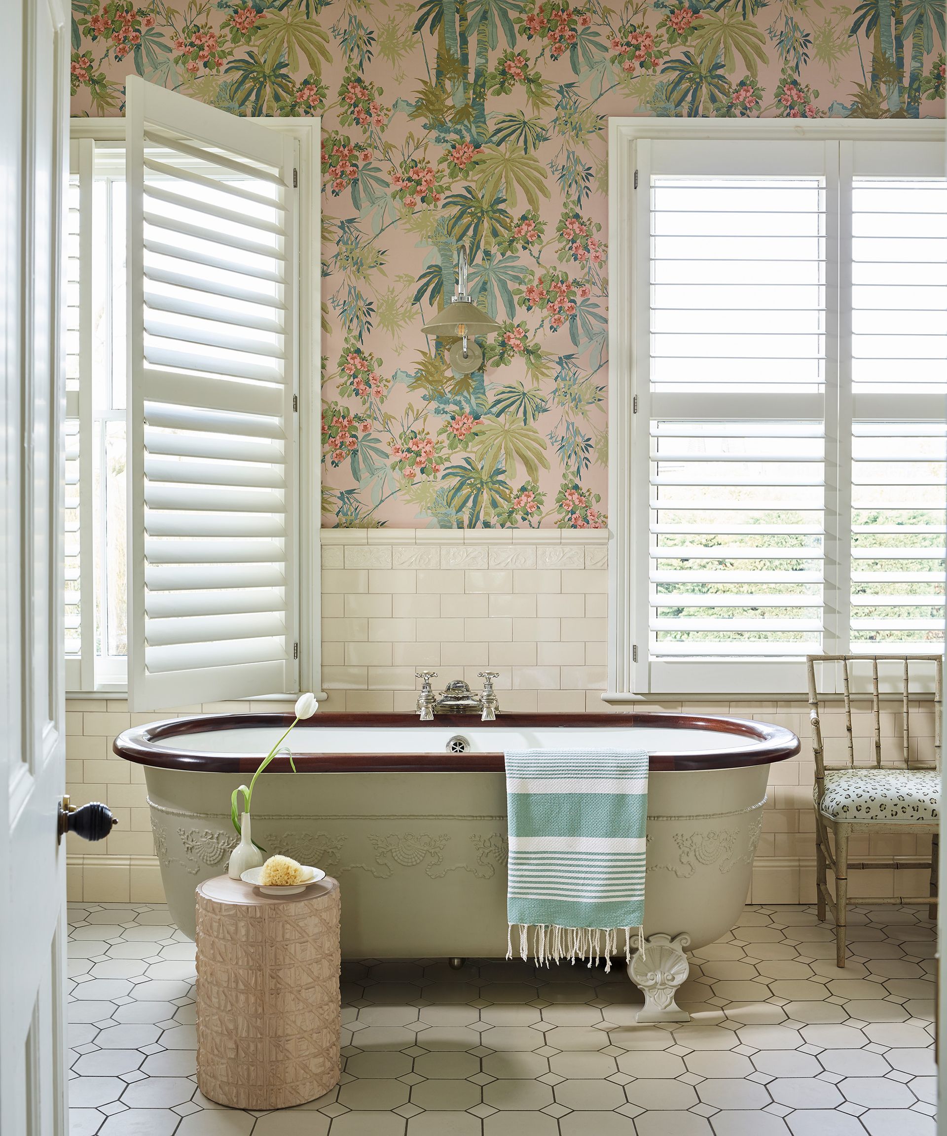 A neutral bathroom with colorful floral and botanical wallpaper above a light green freestanding bath