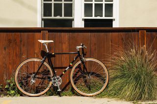 A drivetrain-side image of the State Bicycle Co 4130 Road+ Bob Marley edition black bike leaning against a solid wooden fence.