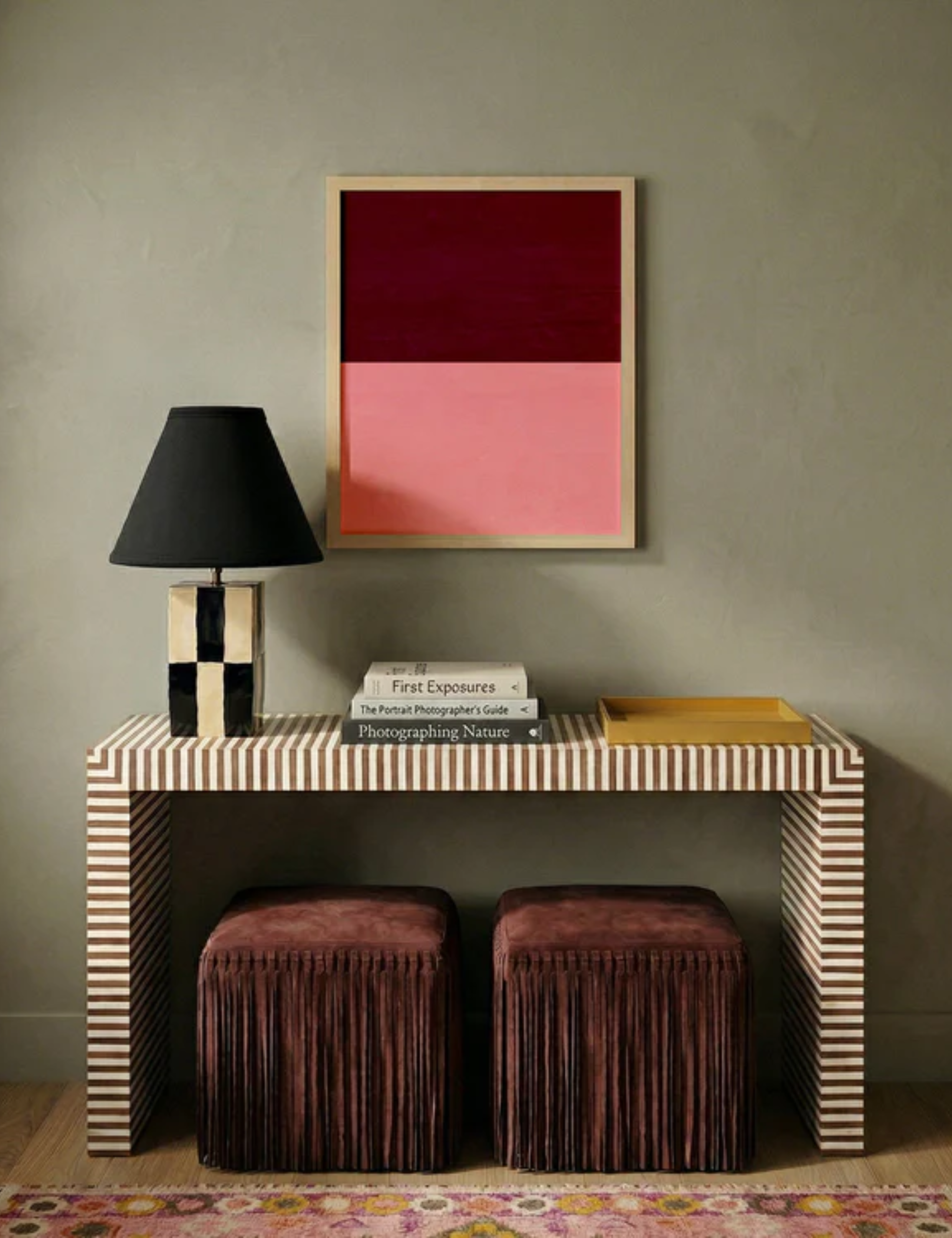 Burgundy-and-pink wall art print styled over a striped console table with a pair of fringe suede ottomans and a checkered table lamp