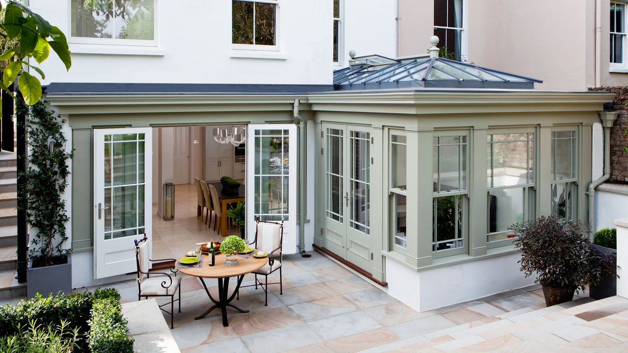 an elegant gray and white orangery addition to a period home by Westbury, with a dining table seen through the open doors and a dining set on the patio