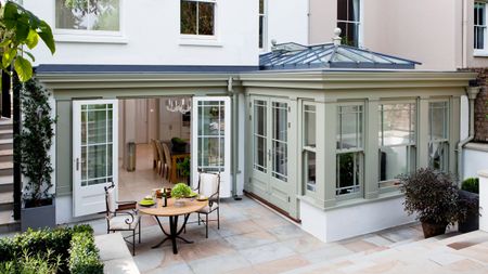 an elegant gray and white orangery addition to a period home by Westbury, with a dining table seen through the open doors and a dining set on the patio