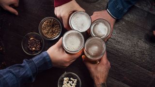 close up on four people's hands holding beers as they raise a toast over a pub table with some small bowls of nuts placed on it