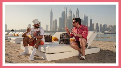 Two cast members from Dubai Bling sitting outside in front of a cityscape of Dubai. Dubai Bling - Production Still Image