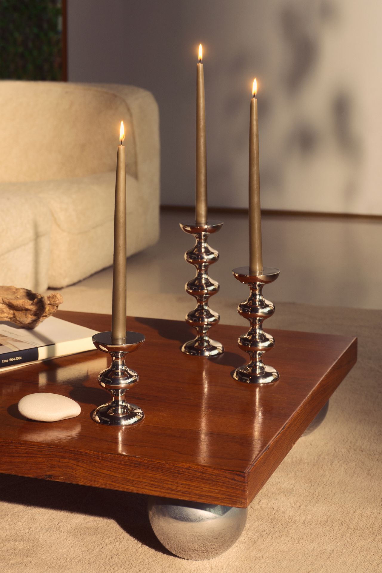 Image of the corner of a wooden coffee table that has round chrome feet. Three abstract, chrome candle holders on the table with beige candles in them.