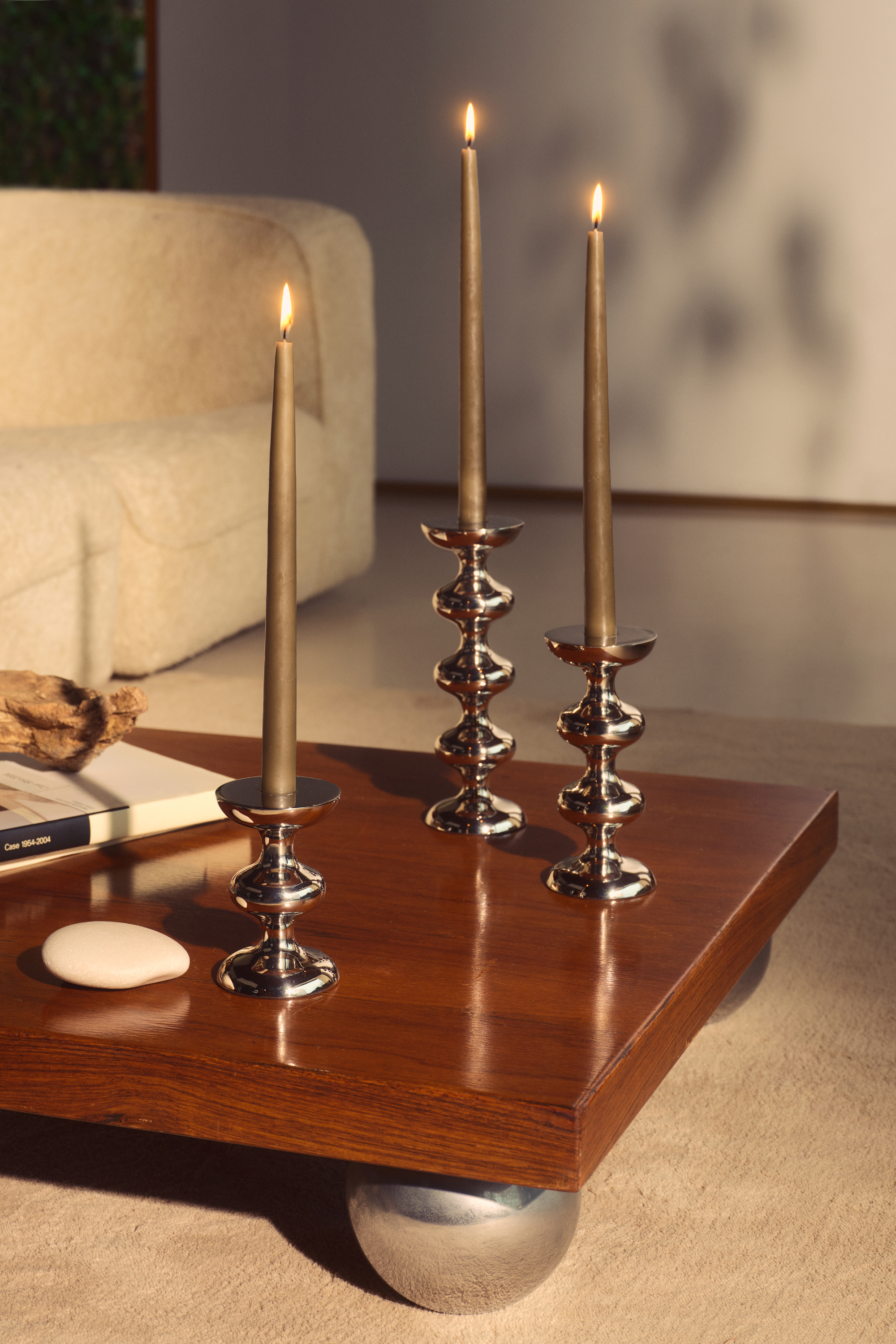 Image of the corner of a wooden coffee table that has round chrome feet. Three abstract, chrome candle holders on the table with beige candles in them. 