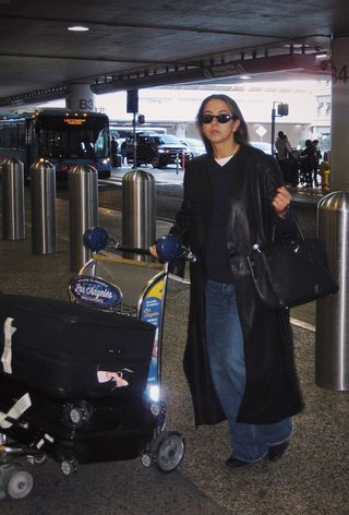 A winter travel outfit is shown in a photo of a woman standing in an airport terminal wearing a black leather trench coat over a white t-shirt layered under a black v-neck sweater that's styled with wide-leg jeans, black ankle boots, sunglasses, and a black Hermes Birkin bag