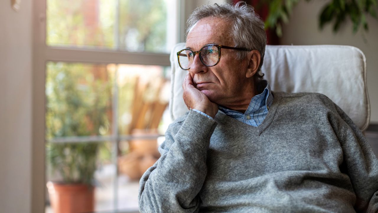 An older man sits beside a window and looks outside, looking a bit nervous.