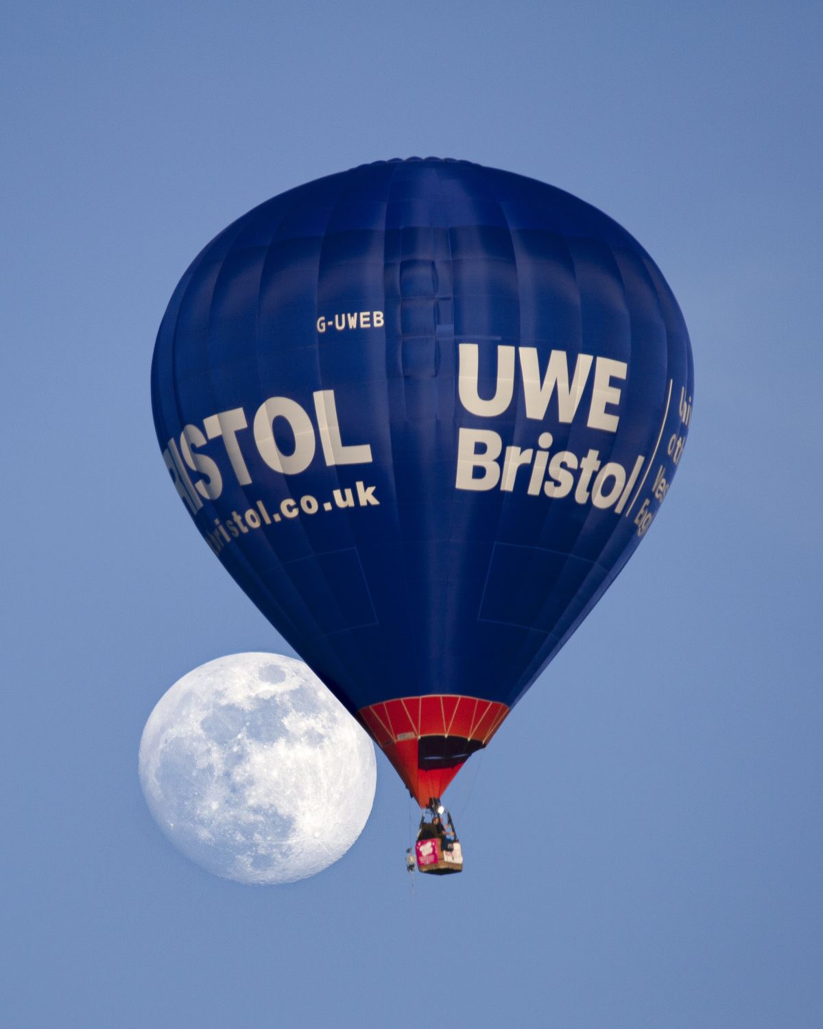 a fully illuminated moon to the left of a hot air balloon against a blue sky.