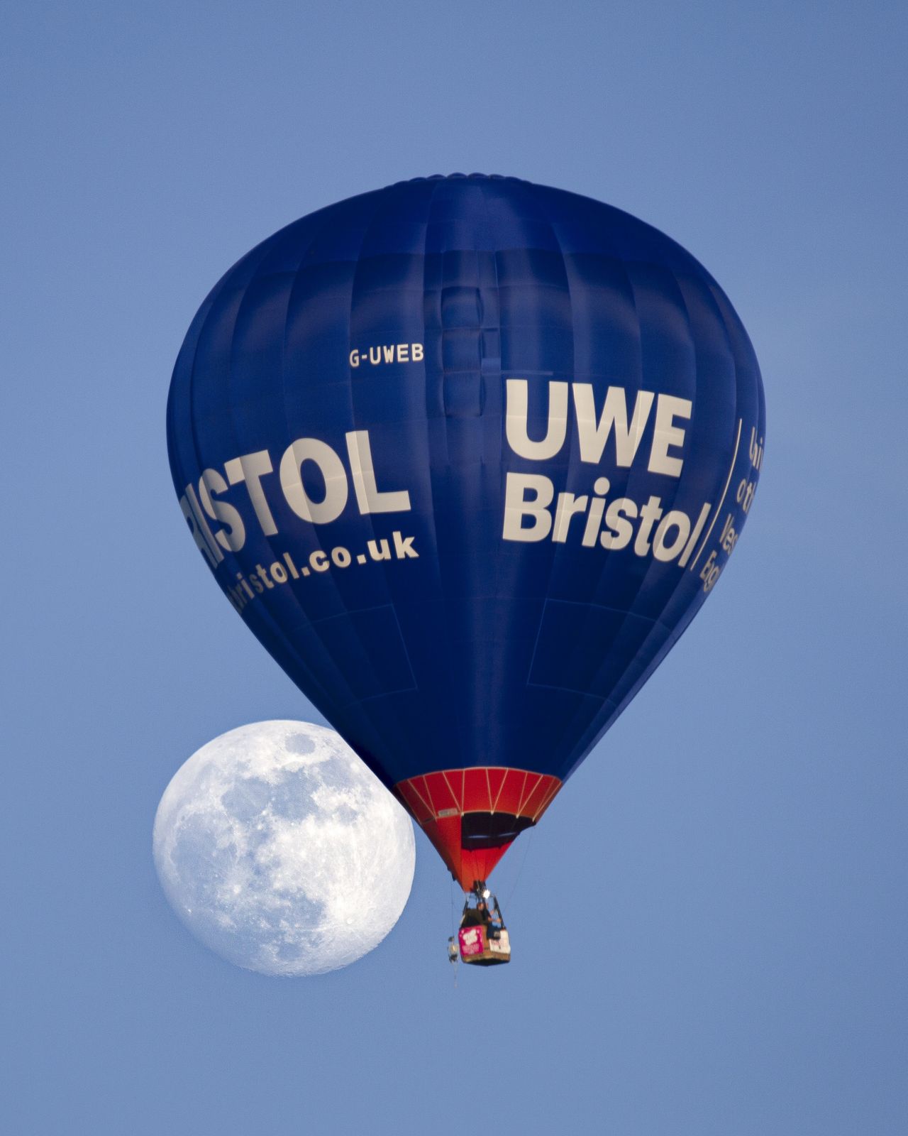 a fully illuminated moon to the left of a hot air balloon against a blue sky.