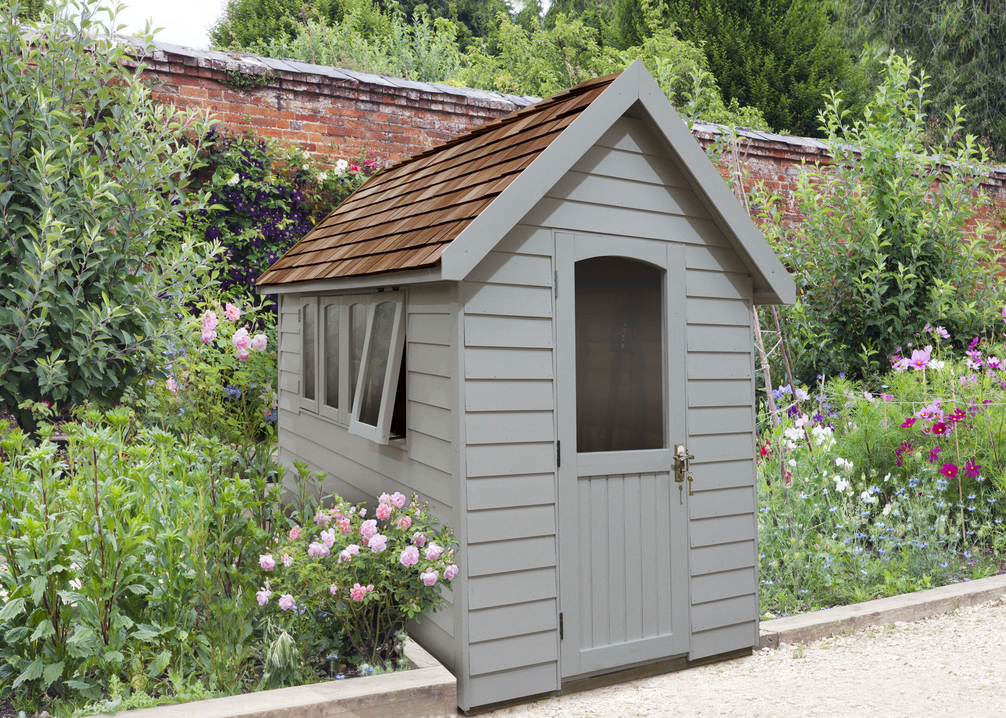 garden shed in sage green with open window, positioned in flower bed with pretty blooms and a brick garden wall behind