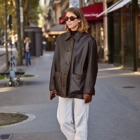 Fashion influencer @monicaainleydlv wearing a chic outfit styled with a brown leather collared coat, cream jeans, leather gloves, and sunglasses.