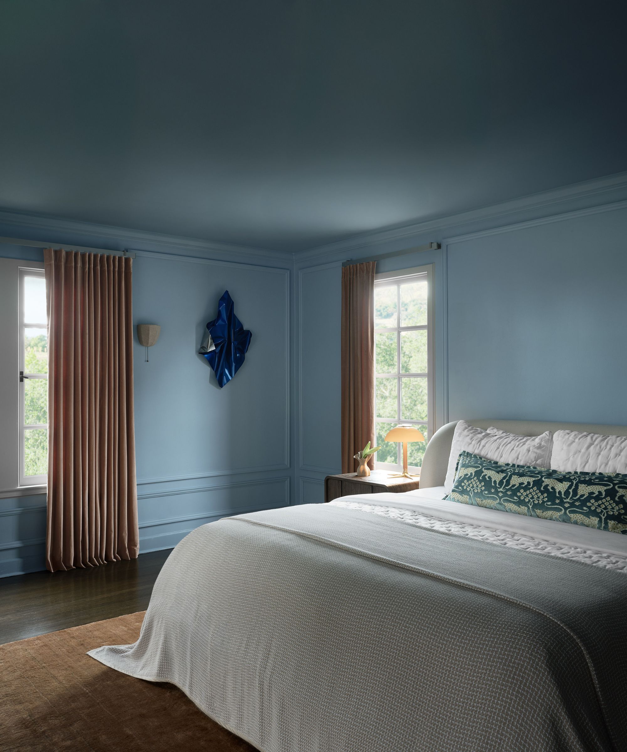 A modern bedroom, color-drenched in mid-blue, with a large bed with soft gray bedding and a long, dark green patterned pillow. Taupe drapes and a caramel-colored rug.