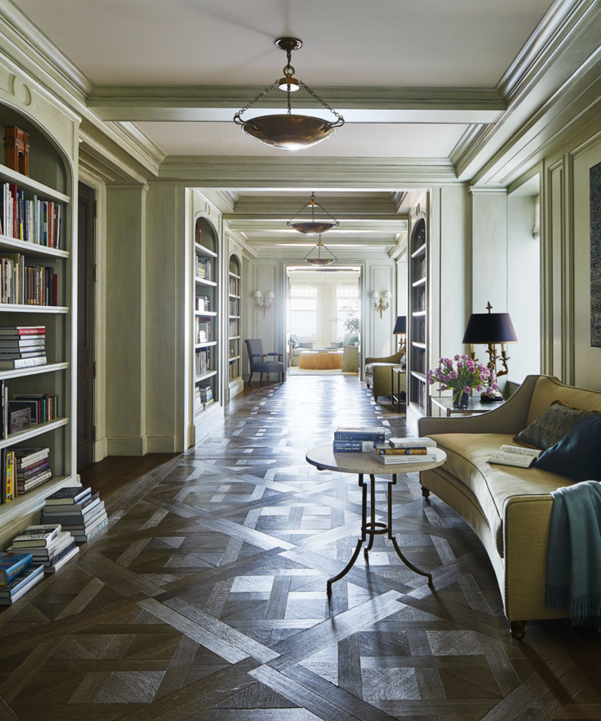 Large, long entryway with bookshelves on either side, wooden flooring, sofa and side table to the lower right hand side of the image, living room with windows in the background