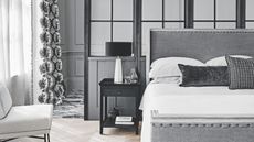 A gray bedroom with a large gray upholstered bed to the right, with white bedding and gray cushions. Beside the bedside table, to the left, is a black bedside table with books, vases, and a white lamp with a black lampshade. To the far right is a sheer white curtain over a window, and a white chair. The walls are gray paneled.