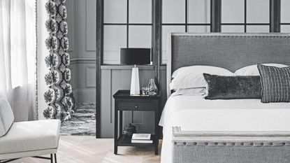 A gray bedroom with a large gray upholstered bed to the right, with white bedding and gray cushions. Beside the bedside table, to the left, is a black bedside table with books, vases, and a white lamp with a black lampshade. To the far right is a sheer white curtain over a window, and a white chair. The walls are gray paneled.