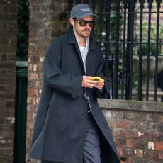 Harry Styles wears black leather loafers with white socks, black straight-leg trousers, a black overcoat and blue button down shirt as he walks through the streets of London.