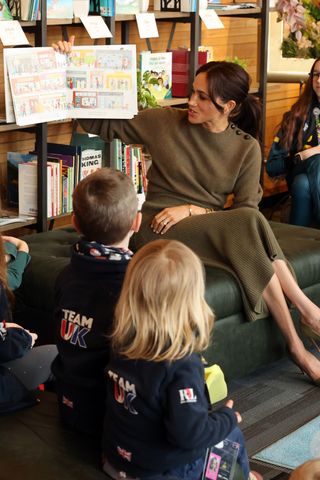 Meghan Markle wearing an olive green sweater and skirt while holding up a story book to a group of children