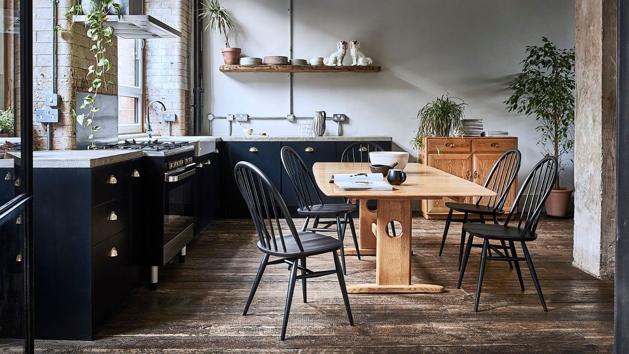 A wooden Ercol dining table with black chairs in a dark kitchen with black cabinets and white countertops 