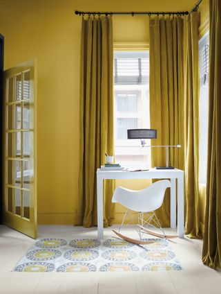 home office with white desk and chair, patterned rug, white flooring, ochre walls and velvet drapes, black desk lamp
