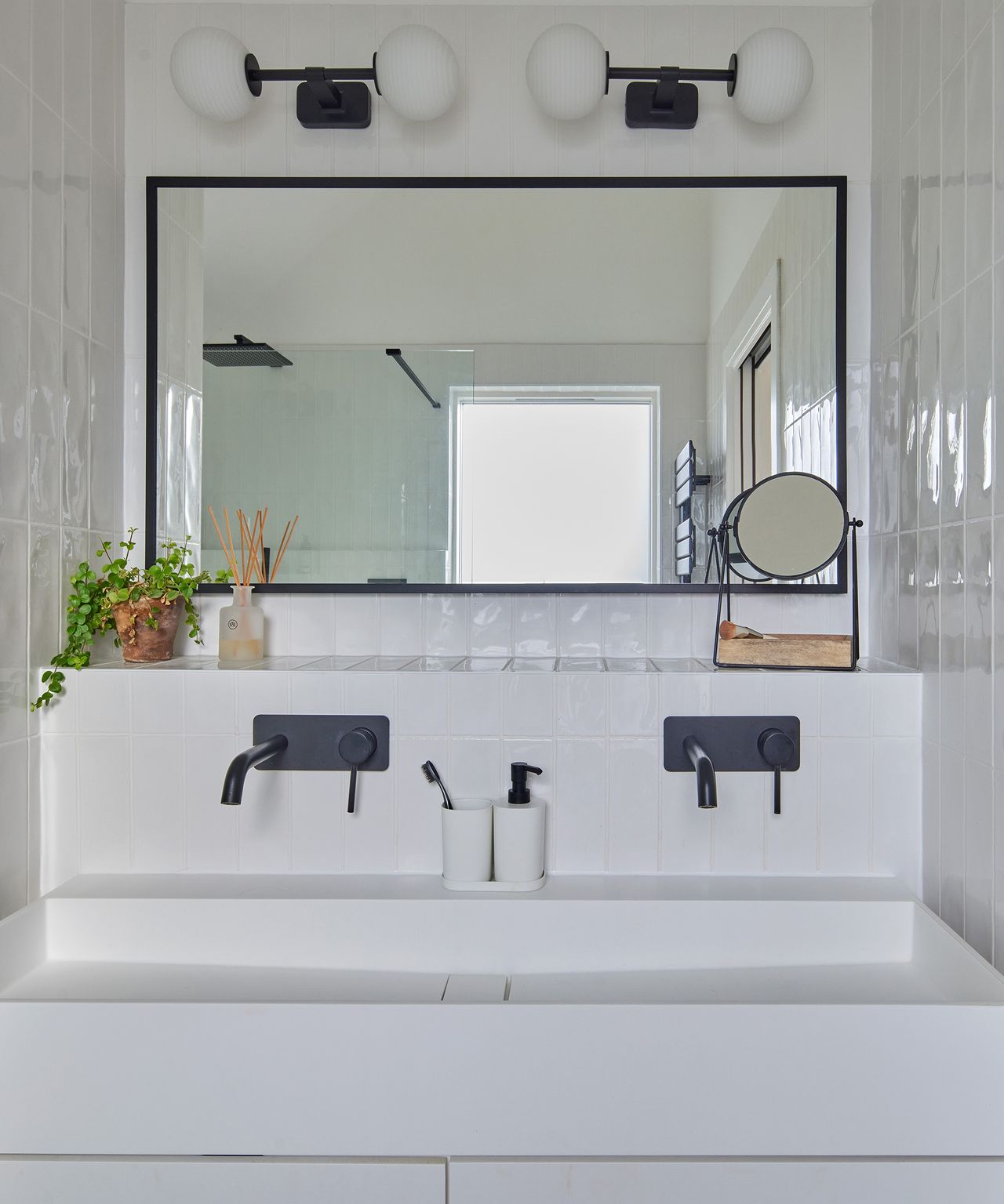 His and hers double sink in a bathroom with black taps and a black frame mirror. There are black wall-mounted lights above the mirror with globe-shaped bulbs