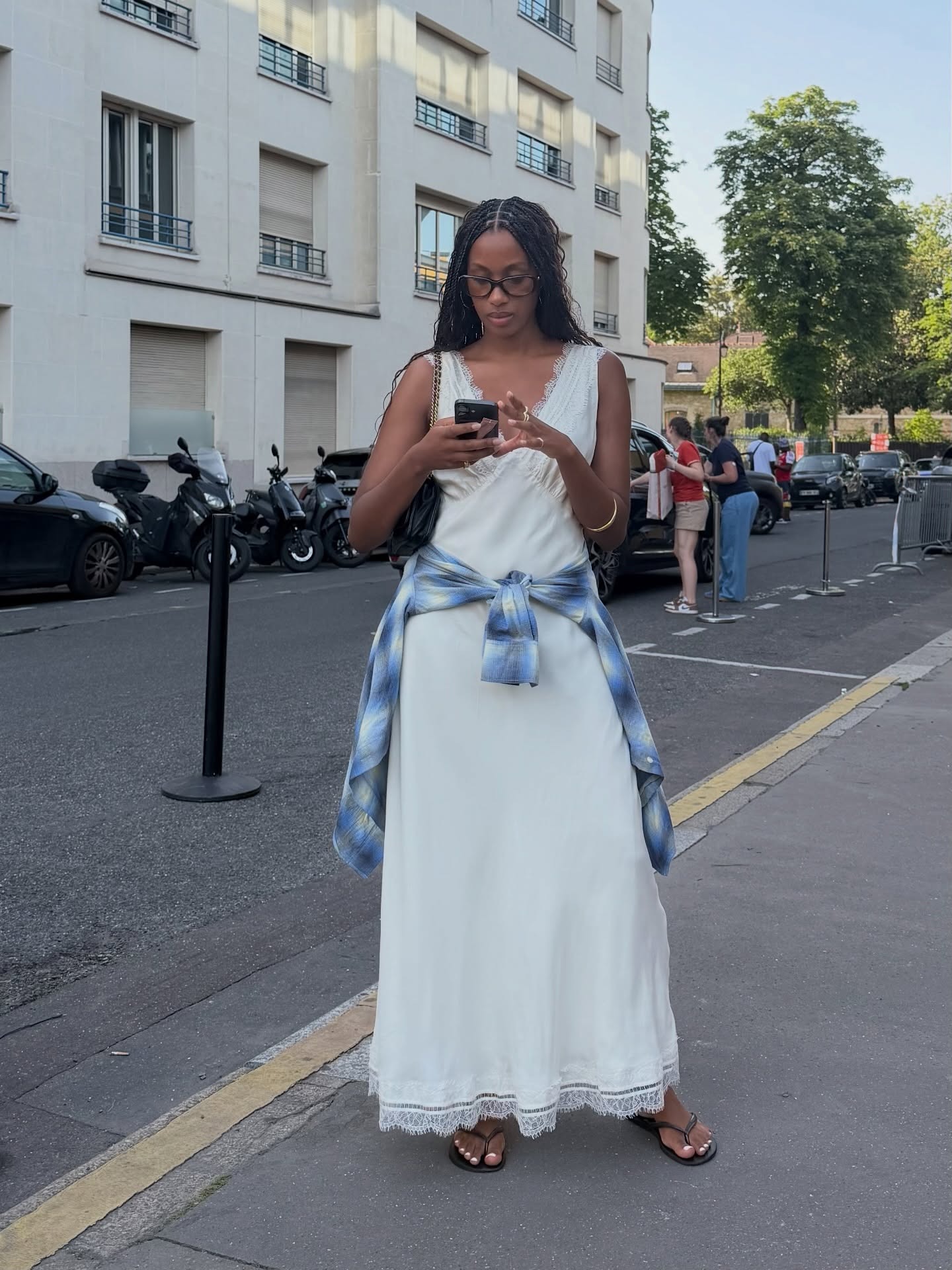 Fashion person amaka.hamelijnck wears june outfit ideas 2026. She wears a white maxi lace trim dress, a flannel shirt, and minimalist black flip-flops. In the recent photo, she is texting while looking down.