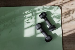 A green yoga matt on the floor with a pair of black dumbells.