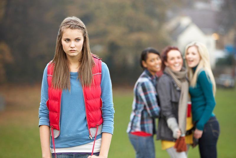 upset teen girl with other girls gossiping in the background