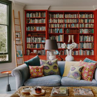 living room paint colours 2023, living room with bright red bookcase, grey sofa, ladder, green painted window frame, leather footstool, books, bright cushions