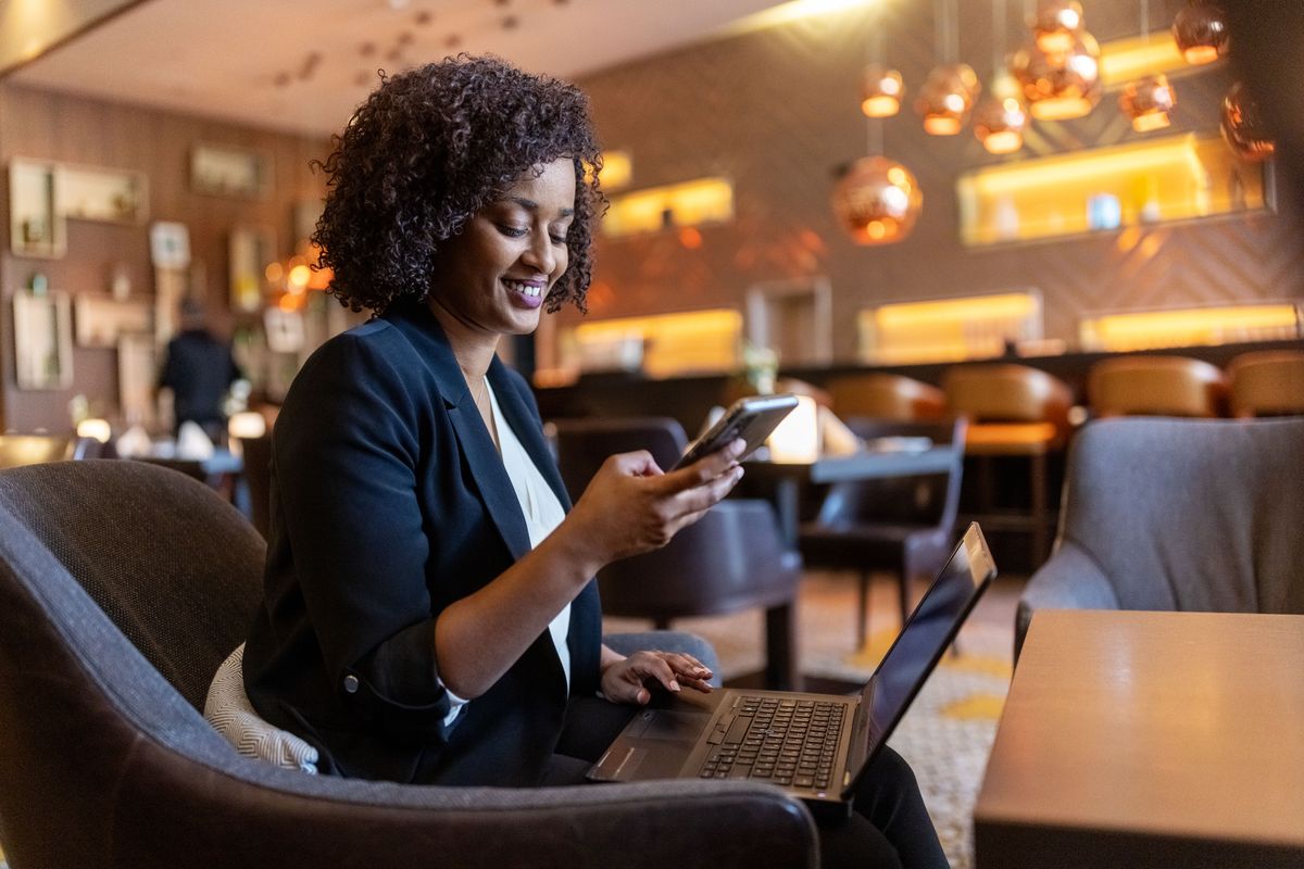 A stock photo of a woman in a public setting working on a mobile phone and a laptop.