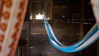 An empty blue hammock is hanging inside of a wood cabin with a palm roof