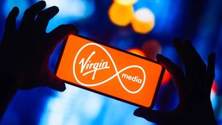 A mobile phone with the Virgin Media logo on the screen is held up in someone's hands front of some blue lights