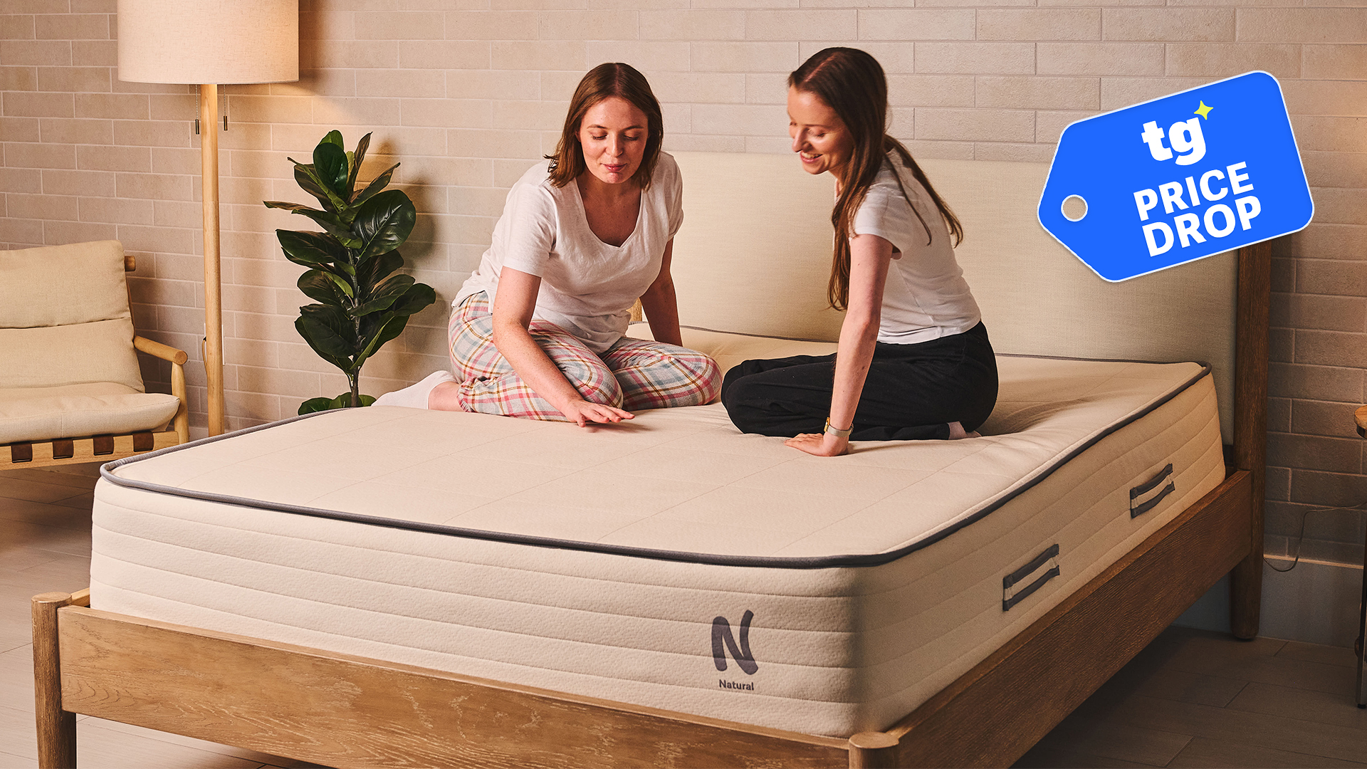 Two women sit on the Nolah Natural 11 mattress in a bedroom. A Tom's Guide Price Drop deals graphic in the top right corner