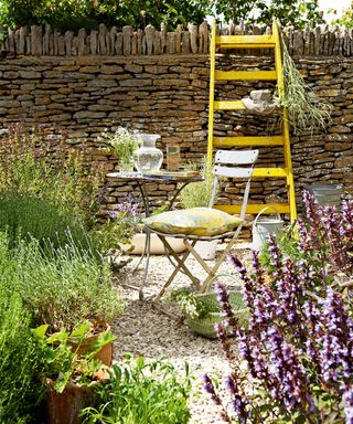 A garden table and chair on gravel in front of a rustic stone wall with a yellow ladder