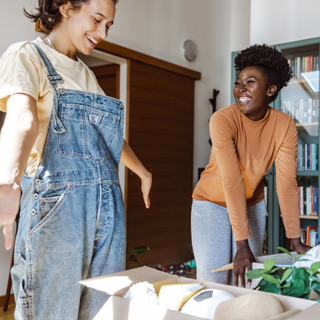 Two female roommates in a new apartment. They are stood behind boxes full of plants and smiling at each other