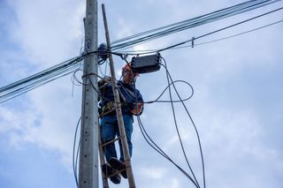 A telecoms worker is shown working from a utility pole ladder while wearing high visibility personal safety clothing, PPE, and a hard hat