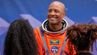 A man in an orange space suit smiles at two people, whose back of their heads are unfocused in the forground on left and right.