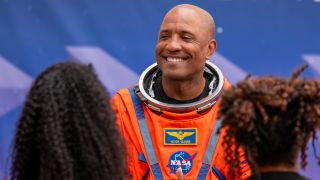 A man in an orange space suit smiles at two people, whose back of their heads are unfocused in the forground on left and right.