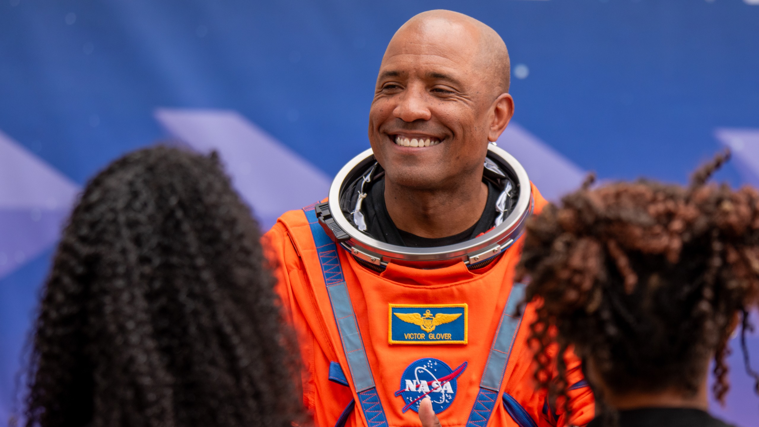 A man in an orange space suit smiles at two people, whose back of their heads are unfocused in the forground on left and right.