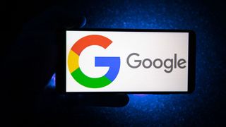 The Google logo shown on a landscape phone, held by a hand in silhouette against a dark blue background